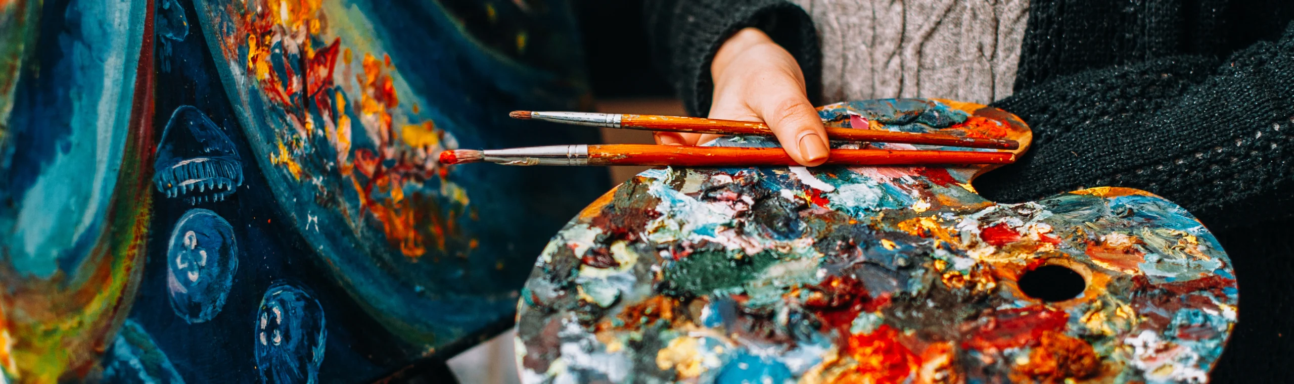 A person holds a paint palette covered in various colors of paint and three paintbrushes. An abstract, colorful painting is visible in the background. The person wears a dark sweater.